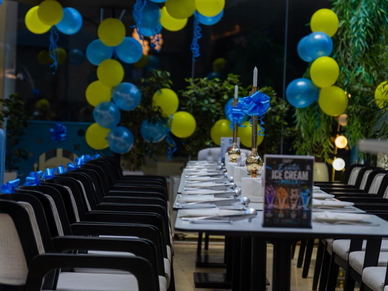 A long table set for a celebration at Robot Cafe Lana Plaza, with blue and yellow balloons and golden candle stands